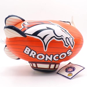 May include: An orange blimp-shaped toy featuring the Denver Broncos logo and the word BRONCOS in white lettering. The toy has dark blue fins and a base, with thin black horizontal lines across the surface and an attached NFL product tag.