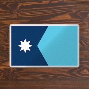 May include: Rectangular magnet featuring a stylized flag design. The flag has a dark blue field with a white eight-pointed star in the upper left corner. A light blue triangle shape extends from the right side.