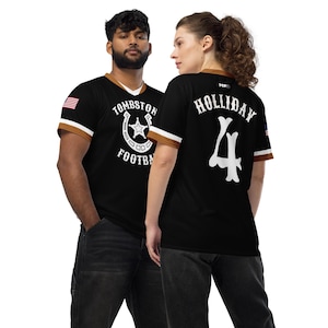 May include: Black and white baseball jersey with a brown trim. The front of the jersey has a horseshoe with a star inside and the text "Tombstone Football". The back of the jersey has the text "Holliday" and the number "4" in a bone design.