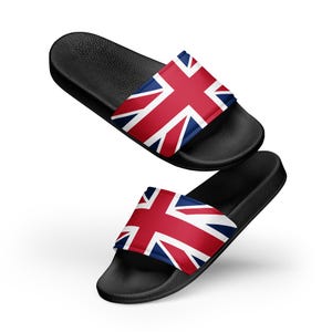 Puede incluir: Dos sandalias negras con el diseño de la bandera de la Union Jack en rojo, blanco y azul. Las sandalias tienen una plantilla texturizada y una correa ancha en la parte superior con la impresión de la bandera. Presentadas sobre fondo blanco.