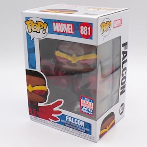 Puede incluir: Figura bobblehead Funko Pop! Marvel Falcon en su caja original. La caja presenta el logotipo de Marvel, el número 881 y el texto "2021 Summer Convention Limited Edition". La figura es roja, marrón y amarilla.