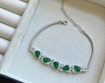 Op de afbeelding: Een zilveren armband met een rij levendige groene, peervormige edelstenen. De armband is bezet met kleine, heldere edelstenen en wordt gepresenteerd in een witte sieradendoos.