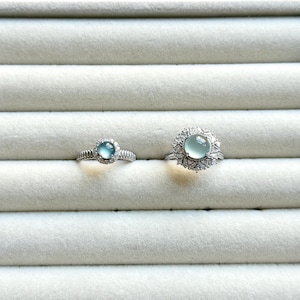 May include: Two silver rings with light blue gemstones. The ring on the left has a smaller gemstone surrounded by a halo of smaller stones. The ring on the right has a larger, round gemstone with a halo of surrounding stones.