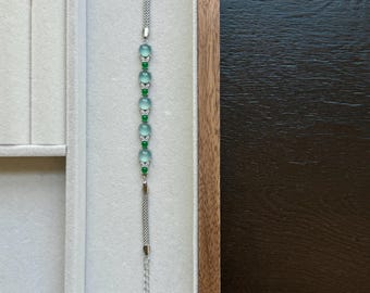 Op de afbeelding: Zilveren armband met lichtblauwe en groene kralen. De armband wordt gepresenteerd in een houten sieradendoos met een wit interieur, tegen een donkere houten achtergrond. De lengte is verstelbaar.