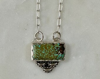 May include: A silver chain necklace with a rectangular turquoise stone pendant. The pendant has a scalloped edge and a silver design below the stone.