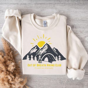 Pode incluir: Uma camisa de moletom cor creme com um gráfico de uma cordilheira, uma fogueira e uma barraca. O texto "Out of Breath Hiking Club" é impresso abaixo do gráfico, com o texto menor "I'll catch up...eventually" embaixo.