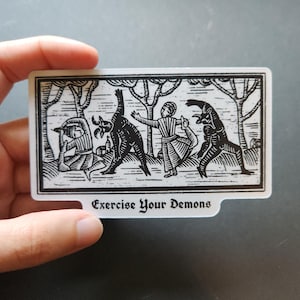 May include: A rectangular sticker featuring a black and white woodcut-style illustration of two people and two horned figures performing yoga poses in a forest setting. The text "Exercise Your Demons" is printed in a gothic font at the bottom.