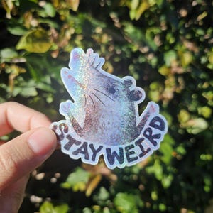 Puede incluir: Una pegatina hologr&aacute;fica con una rata estilizada y las palabras "STAY WEIRD" en negro. La pegatina tiene un acabado brillante e iridiscente. Se muestra sobre un fondo de follaje verde.