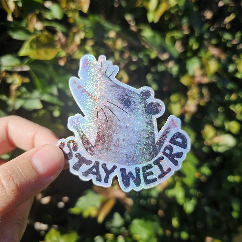 Puede incluir: Una pegatina hologr&aacute;fica con una rata estilizada y las palabras "STAY WEIRD" en negro. La pegatina tiene un acabado brillante e iridiscente. Se muestra sobre un fondo de follaje verde.
