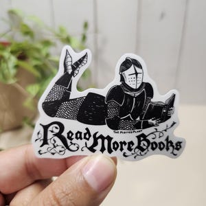 May include: A die-cut sticker featuring a black and white illustration of a knight in full armour lying down and reading a book. The text "Read More Books" is written in a decorative black font below the knight, with "THE PLOTTED PLANT" printed in small letters.