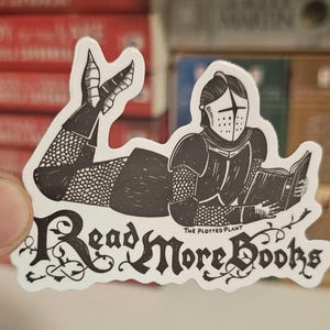 May include: A die-cut sticker featuring a black and white illustration of a knight in full armour lying on their stomach and reading an open book. The text "Read More Books" is written in a decorative font below, with "THE PLOTTED PLANT" printed.