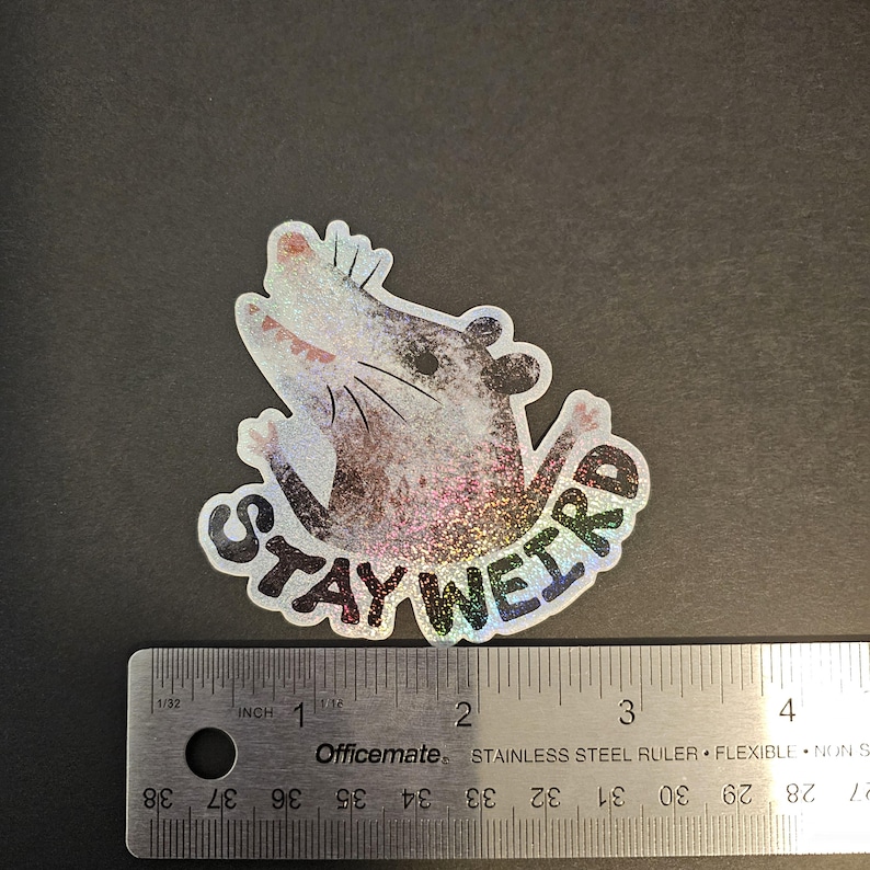 Puede incluir: Una pegatina hologr&aacute;fica con un dibujo de una zarig&uuml;eya y el texto "STAY WEIRD". La zarig&uuml;eya es gris, blanca y rosa con un efecto brillante. Una regla est&aacute; al lado de la pegatina.