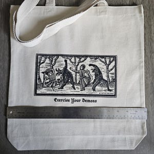 May include: Cream-colored tote bag with a black and white illustration of figures and demons in a forest scene. The text "Exercise Your Demons" is printed below the image. The bag has a canvas strap and is approximately 15 inches wide.