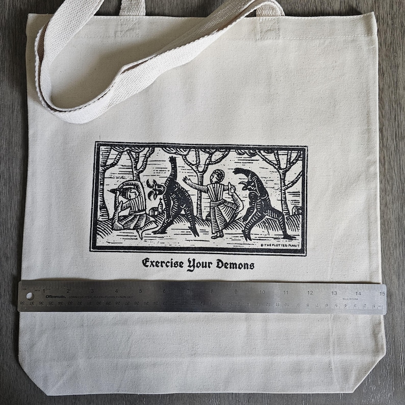 May include: Cream-colored tote bag with a black and white illustration of figures and demons in a forest scene. The text "Exercise Your Demons" is printed below the image. The bag has a canvas strap and is approximately 15 inches wide.