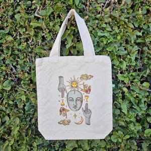 May include: Cream-colored tote bag with a celestial design. The graphic includes a sun, moon, clouds, hands, and a face. The bag has two cream-colored handles and is made of a sturdy fabric.