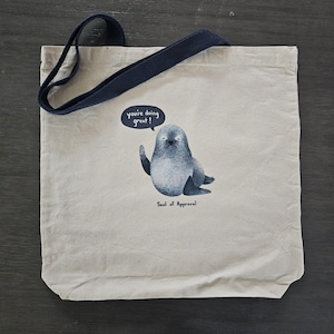 May include: A canvas tote bag with a navy blue handle. The bag features a cartoon seal giving a thumbs up with the text "you're doing great!" in a speech bubble. Below the seal is the text "Seal of Approval."