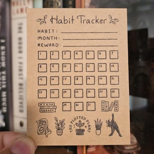 May include: A small, rectangular kraft paper habit tracker card. The card features the text "Habit Tracker" at the top, with spaces for habit, month, and reward. A grid of boxes numbered 1-31 is below. The bottom has plant-themed illustrations and the text "The Plotted Plant."