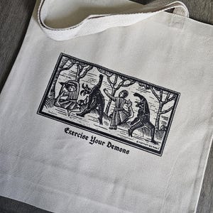 May include: Cream-colored tote bag with a black and white graphic print. The print depicts figures in a forest setting, with the text "Exercise Your Demons" below. The bag has a matching cream-colored handle.