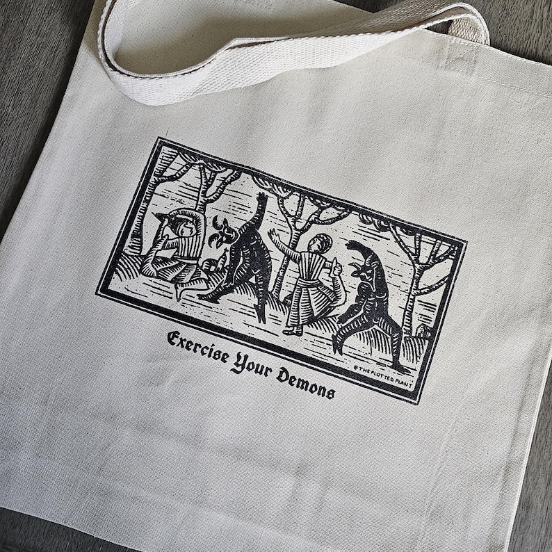 May include: Cream-colored tote bag with a black and white graphic print. The print depicts figures in a forest setting, with the text "Exercise Your Demons" below. The bag has a matching cream-colored handle.