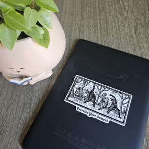 May include: A black e-reader with a sticker featuring a black and white illustration of figures in a forest, with the text "Exercise Your Demons". A small, light pink planter with a plant is in the background.