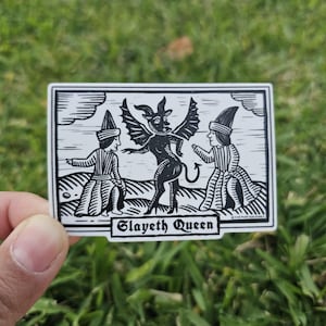 May include: Black and white sticker featuring a stylized illustration of a winged demon and two figures in robes. The text "Slayeth Queen" is displayed in a banner at the bottom of the sticker.