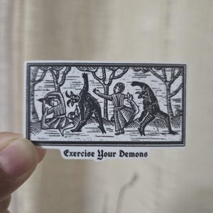 May include: A black and white sticker featuring a stylized illustration of figures interacting with demonic creatures in a forest setting. The text "Exercise Your Demons" is printed at the bottom of the sticker.