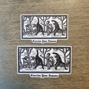 May include: Two black and white stickers with a medieval-style illustration of figures interacting with demonic creatures in a forest setting. The text "Exercise Your Demons" is printed below the image.