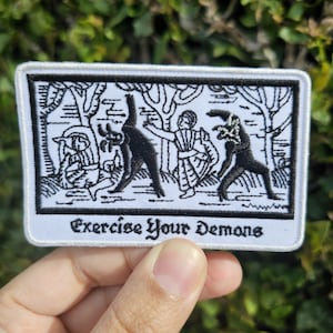 May include: A rectangular white patch featuring a black and white illustration of figures and demons in a forest. The text "Exercise Your Demons" is printed in a stylized font at the bottom of the patch.