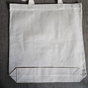 May include: A blank, off-white canvas tote bag with sturdy handles. The bag is rectangular with a flat bottom, designed for carrying items. The material appears to be a durable cotton or similar fabric, suitable for various uses.