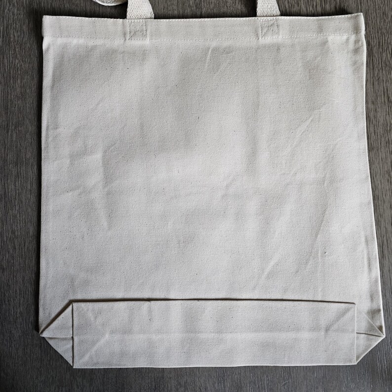 May include: A blank, off-white canvas tote bag with sturdy handles. The bag is rectangular with a flat bottom, designed for carrying items. The material appears to be a durable cotton or similar fabric, suitable for various uses.