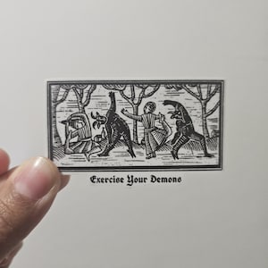 May include: A black and white sticker with a woodcut-style illustration. The image depicts figures interacting with demonic creatures in a forest setting. The text "Exercise Your Demons" is at the bottom.