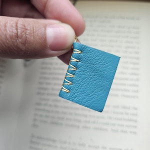 Op de afbeelding: Een kleine, teal-kleurige leren boekenbedel met gouden stiksels. Het miniatuurboekje wordt vastgehouden door een goudkleurige haak. De bedel is een handgemaakt accessoire.