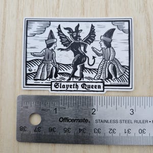 May include: Black and white sticker featuring a stylized illustration of a horned demon with bat wings, flanked by two figures in medieval attire. The text "Slayeth Queen" is displayed below the central image. The sticker is approximately 3 inches long.
