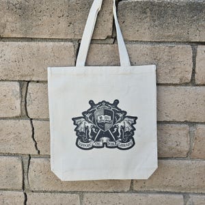 May include: Off-white canvas tote bag with a black and white emblem. The emblem features a shield, dragons, and the text "SUPPORT THE PUBLIC LIBRARY". The bag has two handles and is hanging against a brick wall.