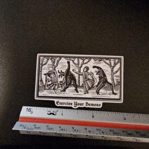 May include: A black and white sticker with the text "Exercise Your Demons." The sticker features a woodcut-style illustration of figures in a forest setting. The sticker is approximately 3.5 inches long.
