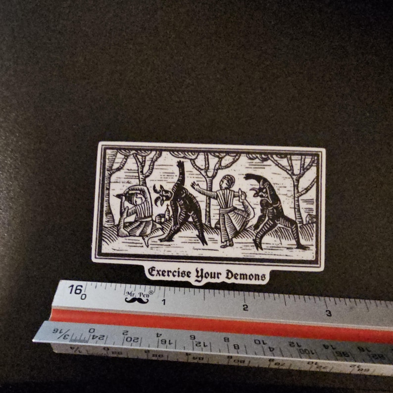 May include: A black and white sticker with the text "Exercise Your Demons." The sticker features a woodcut-style illustration of figures in a forest setting. The sticker is approximately 3.5 inches long.