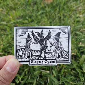 May include: Black and white sticker featuring a central devil-like figure with bat wings, flanked by two figures in medieval attire. The text "Slayeth Queen" is at the bottom. The sticker is held against a background of green grass.