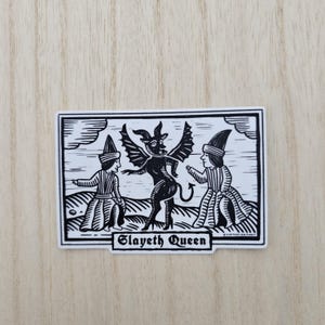 May include: Black and white sticker featuring a devil-like figure with wings, horns, and a tail, flanked by two figures in hats and robes. The text "Slayeth Queen" is displayed below the central image. The sticker has a white border.