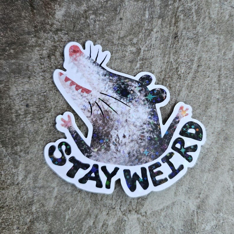 Puede incluir: Una pegatina con un dibujo de una zarig&uuml;eya de dibujos animados con una amplia sonrisa y los brazos extendidos. La zarig&uuml;eya es principalmente gris y blanca con un efecto brillante. Las palabras "STAY WEIRD" est&aacute;n escritas en una fuente curva debajo.