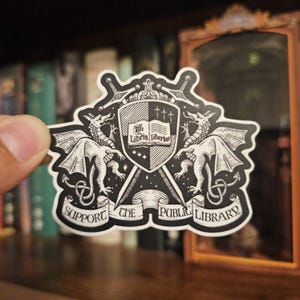 May include: A black and white sticker with a heraldic design. It features a shield with a book and the words "Libri Libertas". Two dragons are on either side, with the text "Support the Public Library" below. The sticker has a decorative border.