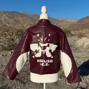 May include: A burgundy and white jacket with a unique design. The jacket features a white graphic on the back and the text "ABOLISH I.C.E." The sleeves are white with burgundy accents. The jacket is displayed on a mannequin.