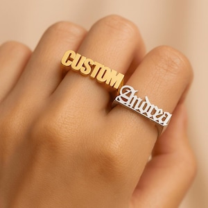 Pode incluir: Dois anéis de nome personalizados. Um anel é dourado com a palavra "CUSTOM" em letras maiúsculas. O outro anel é prateado com o nome "Andrea" em uma fonte ornamentada. Ambos os anéis são usados nos dedos.