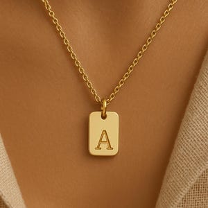 May include: Gold-colored necklace with a rectangular pendant featuring the letter "A". The pendant hangs from a delicate chain. The necklace is displayed against a neutral background.