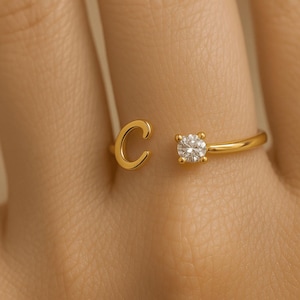 May include: A gold-coloured open ring with the letter "C" and a sparkling, round-cut diamond. The ring is set on a finger, highlighting the delicate design and the brilliance of the gemstone. The ring's band is thin and elegant.