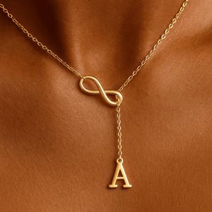 May include: A gold-colored necklace featuring an infinity symbol and a dangling letter "A". The chain is delicate, and the pendant is centered. The necklace is worn against a brown skin tone.