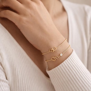 Op de afbeelding: Twee goudkleurige armbanden. De bovenste armband heeft een hartvormig ontwerp met de letters "AC". De onderste armband heeft een hartvormig ontwerp met de letters "On" en een kleine, heldere edelsteen. De armbanden zitten om de pols van een persoon.