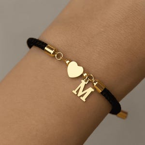 May include: A black cord bracelet with gold-coloured details. It features a gold heart charm and a gold letter "M" charm. The bracelet is designed to be worn on the wrist.