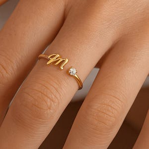 Peut inclure: Une bague dorée avec la lettre "M" en écriture cursive. La bague est ornée d'une petite pierre précieuse transparente. La bague est portée au doigt.