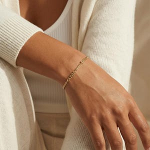 May include: A gold bracelet with the names "OLIVIA" and "EMILY" inscribed. The bracelet is a fine chain design, worn on a wrist. The background is a soft, neutral tone.