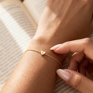 May include: A delicate gold bracelet with a heart-shaped charm. The charm features the letter "B" in the center. The bracelet is on a wrist, with a book in the background.